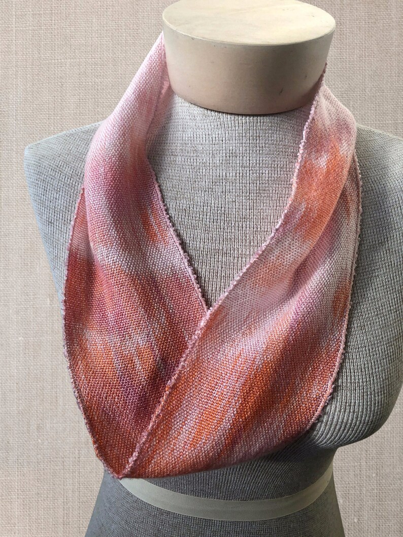 May include: A pink and orange woven scarf with a tie-dye pattern. The scarf is displayed on a mannequin.