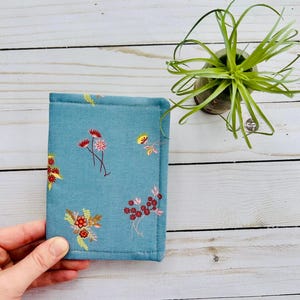 May include: A teal fabric DIY tract holder with a floral pattern in red, yellow, and green. The text "DIY Tract Holder" is in red at the top. A small potted plant is in the background.