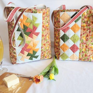 May include: Two quilted tote bags with floral and geometric patterns. One bag features a patchwork design with orange, green, and red shapes. The other has a similar design with diamond shapes. Both have white straps.