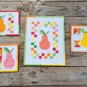 May include: Four small quilted squares with a pear design in the center. Each square has a different color scheme with orange, yellow, green, and red fabrics. The squares are on a wooden background.