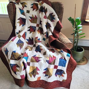 Pode incluir: Um edredom feito à mão com fundo branco e desenhos de patas de urso coloridos e estilizados. O edredom apresenta uma borda vermelha escura e é drapeado sobre uma poltrona estampada. Os blocos de patas de urso do edredom são em tons de azul, amarelo e vermelho.