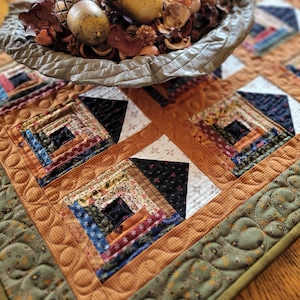May include: A handmade, quilted table runner with a patchwork design. The runner features squares of various fabrics in shades of brown, blue, and red, with a green border. A decorative bowl with dried flowers and eggs sits on top.