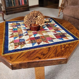 May include: A handcrafted, multi-coloured star quilt, with a blue border, rests on a wooden octagonal table. A small, brown vase with dried flowers sits atop the quilt. The table is made of dark wood.