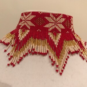May include: A red and white beaded choker necklace with a fringe of gold and white beads. The choker features a geometric pattern of stars.