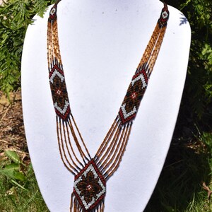 May include: A multi-strand beaded necklace with a geometric pattern in brown, black, white, and red. The necklace has a long, dangling pendant with a similar geometric design.