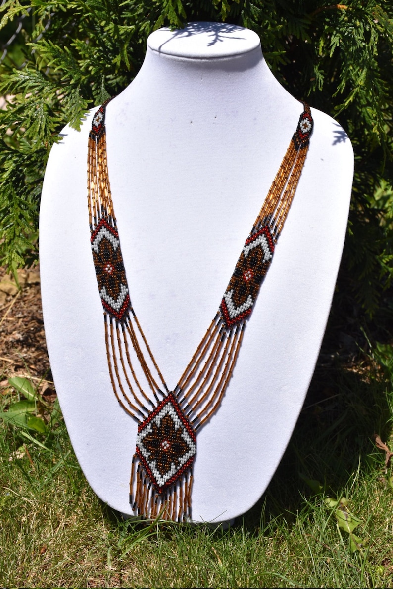 May include: A multi-strand beaded necklace with a geometric pattern in brown, black, white, and red. The necklace has a long, dangling pendant with a similar geometric design.