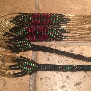 Puede incluir: Un collar de cuentas de m&uacute;ltiples hilos con cuentas rojas, verdes y negras. El collar tiene un patr&oacute;n geom&eacute;trico y est&aacute; hecho con cuentas de color dorado.