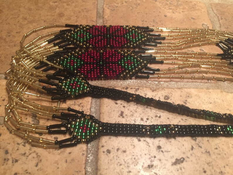 Puede incluir: Un collar de cuentas de m&uacute;ltiples hilos con cuentas rojas, verdes y negras. El collar tiene un patr&oacute;n geom&eacute;trico y est&aacute; hecho con cuentas de color dorado.