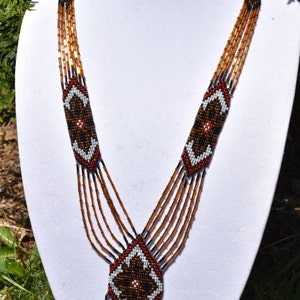 May include: A multi-strand beaded necklace with a geometric pattern in brown, black, white, and red. The necklace has a long, dangling pendant with a similar geometric pattern.