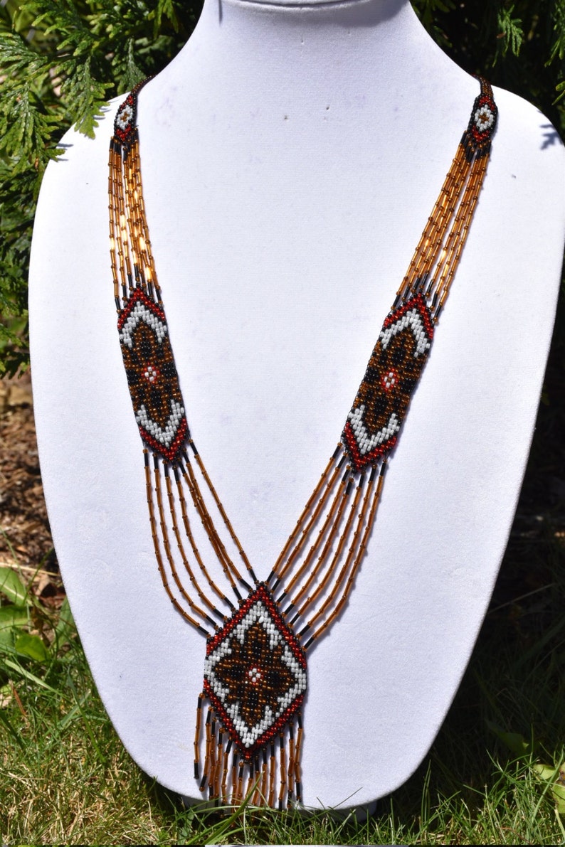 May include: A multi-strand beaded necklace with a geometric pattern in brown, black, white, and red. The necklace has a long, dangling pendant with a similar geometric pattern.