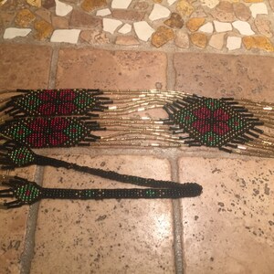 Puede incluir: Un collar largo de cuentas negras, doradas, rojas y verdes. El collar presenta un patr&oacute;n repetitivo de formas geom&eacute;tricas y flores.