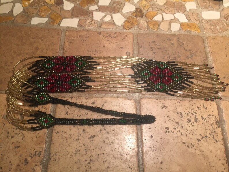 Puede incluir: Un collar largo de cuentas negras, doradas, rojas y verdes. El collar presenta un patr&oacute;n repetitivo de formas geom&eacute;tricas y flores.