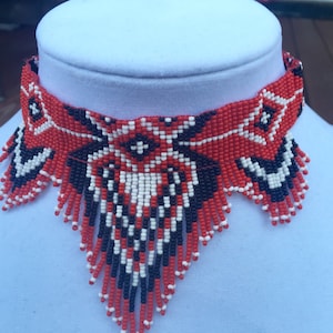 May include: A red, white, and black beaded choker necklace with a geometric pattern. The necklace has a fringe design and is made of small beads.