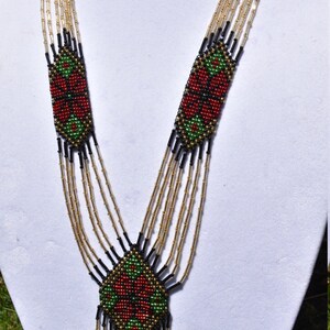 Puede incluir: Un collar de cuentas de m&uacute;ltiples hilos con un dise&ntilde;o geom&eacute;trico que presenta cuentas rojas, verdes y negras. El collar tiene un borla en la parte inferior.