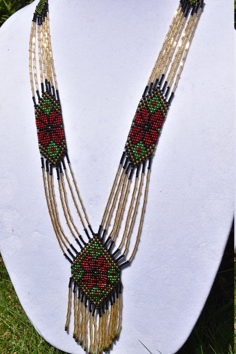 Puede incluir: Un collar de cuentas de m&uacute;ltiples hilos con un dise&ntilde;o geom&eacute;trico que presenta cuentas rojas, verdes y negras. El collar tiene un borla en la parte inferior.