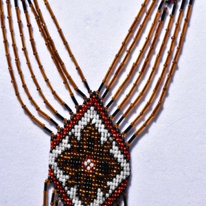 May include: A beaded necklace with a geometric pattern in brown, white, red, and black beads. The necklace has multiple strands of brown beads and a pendant with a diamond shape.