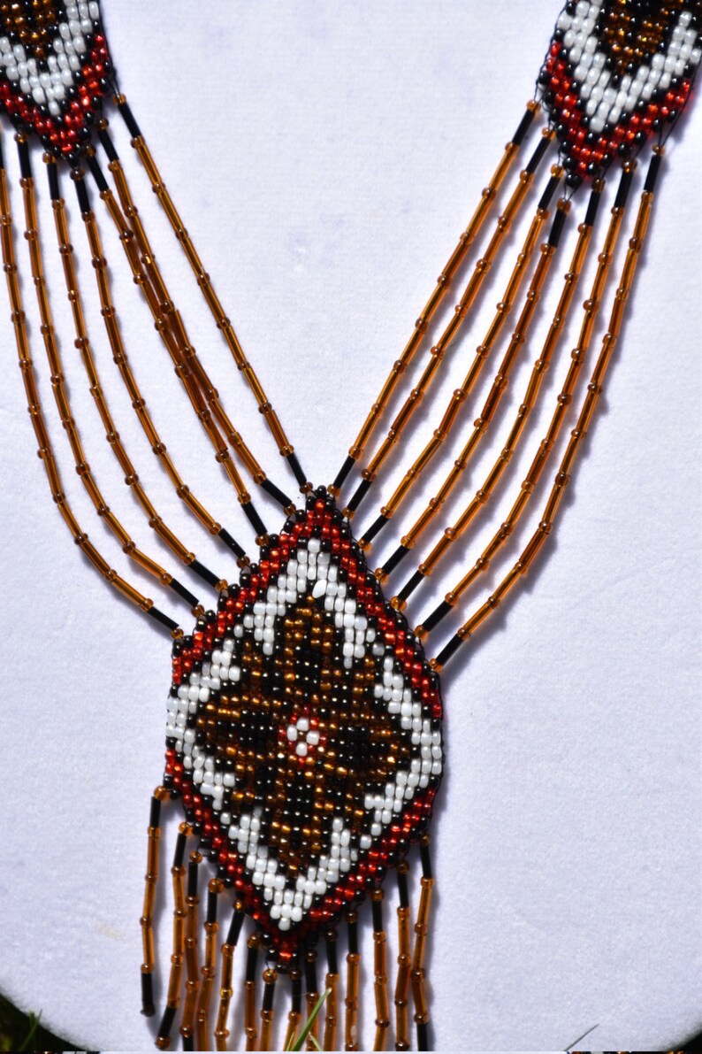 May include: A beaded necklace with a geometric pattern in brown, white, red, and black beads. The necklace has multiple strands of brown beads and a pendant with a diamond shape.