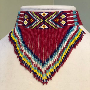 May include: A red beaded choker necklace with a geometric pattern in blue, white, and yellow. The necklace has a fringe of red beads.