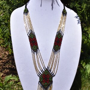 Puede incluir: Un collar de cuentas de m&uacute;ltiples hilos con un dise&ntilde;o geom&eacute;trico en rojo, verde y negro. El collar tiene una borla larga y colgante.