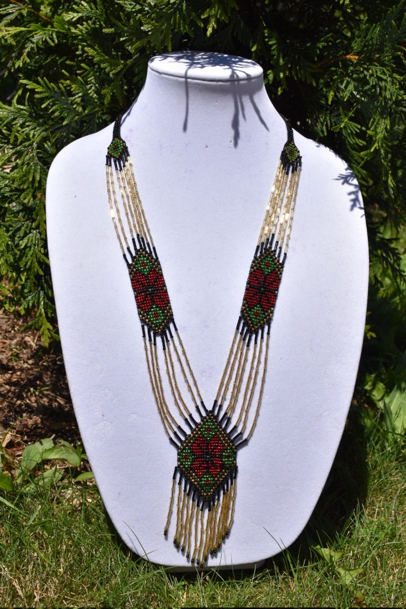 Puede incluir: Un collar de cuentas de m&uacute;ltiples hilos con un dise&ntilde;o geom&eacute;trico en rojo, verde y negro. El collar tiene una borla larga y colgante.
