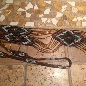May include: A long beaded necklace with a geometric pattern in shades of brown, red, white, and black. The necklace has a fringe of brown beads.