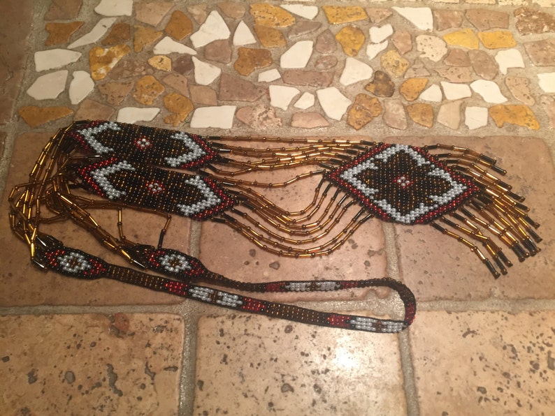 May include: A long beaded necklace with a geometric pattern in shades of brown, red, white, and black. The necklace has a fringe of brown beads.
