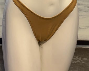 Puede incluir: Una braga de bikini tipo tanga de color tostado sólido con un diseño de pierna de corte alto y una apariencia suave y sin costuras. La prenda se muestra en un torso de maniquí blanco, destacando la construcción minimalista y los bordes cosidos.