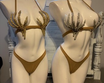 Puede incluir: Dos conjuntos de bikini color canela sobre maniquíes blancos. Cada parte superior presenta un par de manos de esqueleto moldeadas en color beige claro sobre las copas. Las partes inferiores son de corte alto con tiras laterales finas.