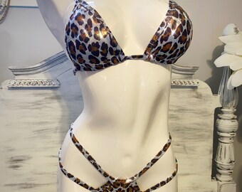Puede incluir: Un conjunto de bikini de dos piezas sobre un maniquí. La parte superior del bikini es de estilo triangular con tirantes finos. La parte inferior tiene un diseño de tiras. Ambas piezas presentan un estampado de leopardo con manchas marrones sobre fondo plateado.