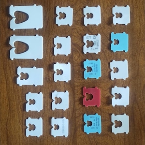 May include: An assortment of white, blue, and red bread bag clips arranged on a wooden surface. Some clips have printed text and numbers. The clips are made of plastic and have a distinctive shape with a heart-shaped opening.