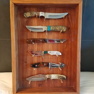 Pode incluir: Uma vitrine de madeira com cinco facas montadas na parede. As facas t&ecirc;m cabos e l&acirc;minas de diferentes cores. A vitrine &eacute; de madeira escura com uma moldura de madeira clara.