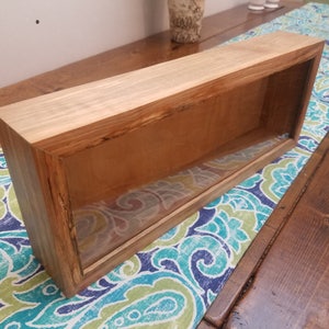 Pode incluir: Uma vitrine retangular de madeira com uma frente de vidro. A vitrine &eacute; feita de madeira marrom claro com um acabamento natural. A vitrine est&aacute; sentada em uma mesa com uma toalha de mesa estampada azul e verde.