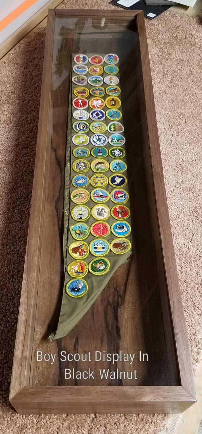 Pode incluir: Uma exibi&ccedil;&atilde;o de distintivos de m&eacute;rito de Boy Scout emoldurada em madeira de noz preta. Os distintivos s&atilde;o exibidos em uma faixa verde.