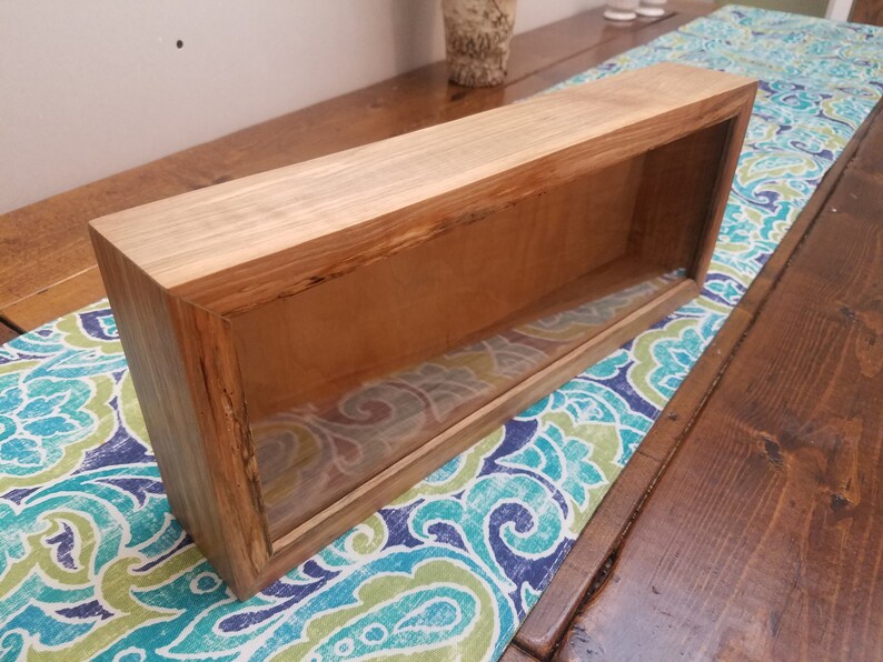 Pode incluir: Uma vitrine retangular de madeira com uma frente de vidro. A vitrine &eacute; feita de madeira marrom claro com um acabamento natural. A vitrine est&aacute; sentada em uma mesa com uma toalha de mesa estampada azul e verde.