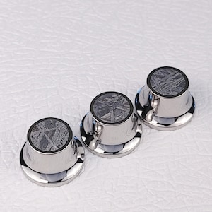 Puede incluir: Tres botones de guitarra plateados con una incrustación de meteorito negro. Los botones son redondos con un borde elevado y una parte superior plana. La incrustación de meteorito es de color gris oscuro con un patrón de remolino.