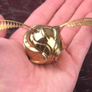 May include: A golden Snitch from the Harry Potter series, held in a hand. The Snitch is a small, spherical object with intricate detailing and two delicate, golden wings extending from its sides. The wings have a detailed, feather-like design.