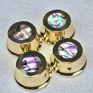 Può includere: Quattro manopole per chitarra color oro con intarsi iridescenti di conchiglia di abalone. Le manopole hanno una parte superiore arrotondata e una base svasata. La conchiglia di abalone mostra colori come viola, verde e blu.