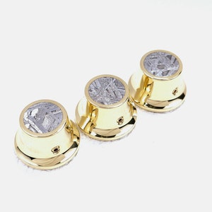 May include: Three gold guitar knobs with a silver meteorite inlay. The knobs are round with a raised edge and a small hole in the centre.