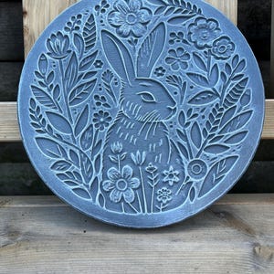 May include: A circular grey stone plaque featuring a relief carving of a rabbit surrounded by detailed floral and leaf patterns. The design includes various blossoms and foliage arranged symmetrically around the central rabbit figure.