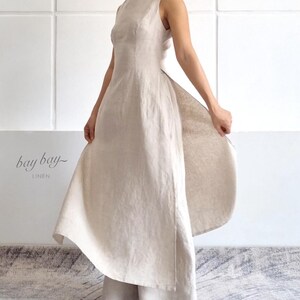 Peut inclure: Une robe sans manches en lin beige avec une encolure haute et un corsage ajusté qui se transforme en une jupe large et fluide avec un panneau latéral ouvert. Le texte "bay bay LINEN" est visible sur le côté gauche en police cursive.