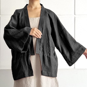 Może przedstawiać: Ciemnoszara lniana kurtka w stylu kimono z szerokimi rękawami i luźnym krojem. Kurtka jest otwarta z przodu i noszona na jasnobeżowej sukience. Tekst "bay bay~ LINEN" jest wydrukowany kursywą po prawej stronie u dołu.
