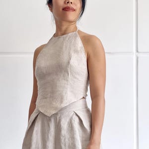 Könnte beinhalten: Ein beiges zweiteiliges Set aus Leinen, bestehend aus einem ärmellosen Neckholder-Top mit spitzem Saum und einer passenden hoch taillierten Hose mit weitem Bein. Der Text bay bay LINEN ist unten links im Bild zu sehen.
