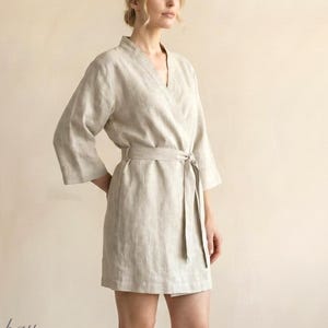 以下が含まれることがあります： ベージュ色のラップ式ローブで、七分袖とウエストに共布の結び紐が付いています。Vネックラインで、丈は太ももの中間まであります。左下隅に「bay bay LINEN」という文字が印刷されています。