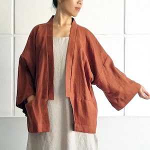 Może przedstawiać: Rdzawy lniany płaszcz kimono z dwiema przednimi kieszeniami, noszony na długiej, jasnobeżowej lnianej sukience. Sukienka ma boczne rozcięcie. Tekst "bay bay~ LINEN" jest widoczny po prawej stronie obrazu.