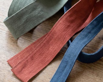 May include: Three long, flat strips of fabric in sage green, terracotta orange, and navy blue. Each strip features visible stitching along the edges and a textured weave. The text "bay bay LINEN" is printed in black near the green fabric strip.