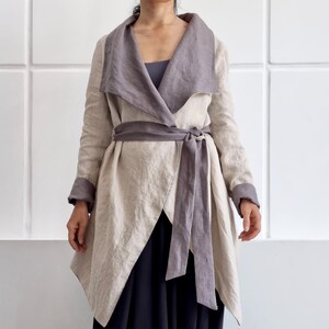 May include: A beige linen jacket with a wide, dark gray shawl collar, long sleeves with dark gray cuffs, and a matching dark gray tie belt at the waist. The jacket is worn over a dark gray top and loose black trousers. The text "bay bay LINEN" is visible.