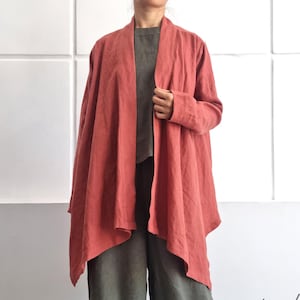 Może przedstawiać: Kobieta ubrana w rdzawy lniany kardigan, szarą górę i oliwkowozielone spodnie z szerokimi nogawkami. Strój ma luźny krój, odpowiedni do noszenia na co dzień. Tło stanowi biała ściana. Widoczny jest tekst "bay bay~ LINEN".