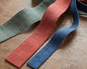 May include: Three long, flat fabric strips in sage green, terracotta orange, and navy blue. Each strip features visible stitching along the edges and a textured, woven appearance. The strips are arranged in overlapping, wavy lines.