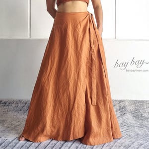 May include: A rust-coloured two-piece set consisting of a cropped camisole top with a button-front closure and a matching floor-length wrap skirt with a side tie. The fabric has a visible linen-like texture. Text reads bay bay baybaylinen.com.