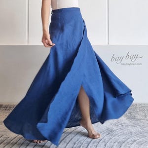Può includere: Una gonna a portafoglio blu intenso lunga fino ai piedi, con vita alta e una silhouette fluida e svasata. La gonna presenta una sovrapposizione frontale prominente che crea un effetto spacco durante il movimento. Testo 'bay bay' e 'baybaylinen.com'.